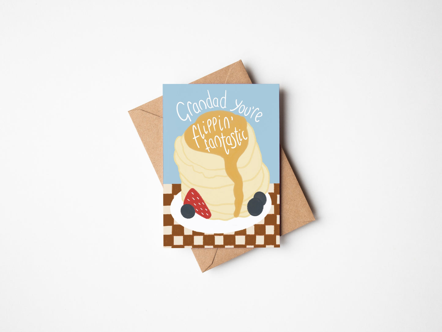 A6 premium textured greeting card with blue background, a stack of pancakes with drizzling syrup, berries and chequered brown tablecloth. Includes brown Kraft envelope.