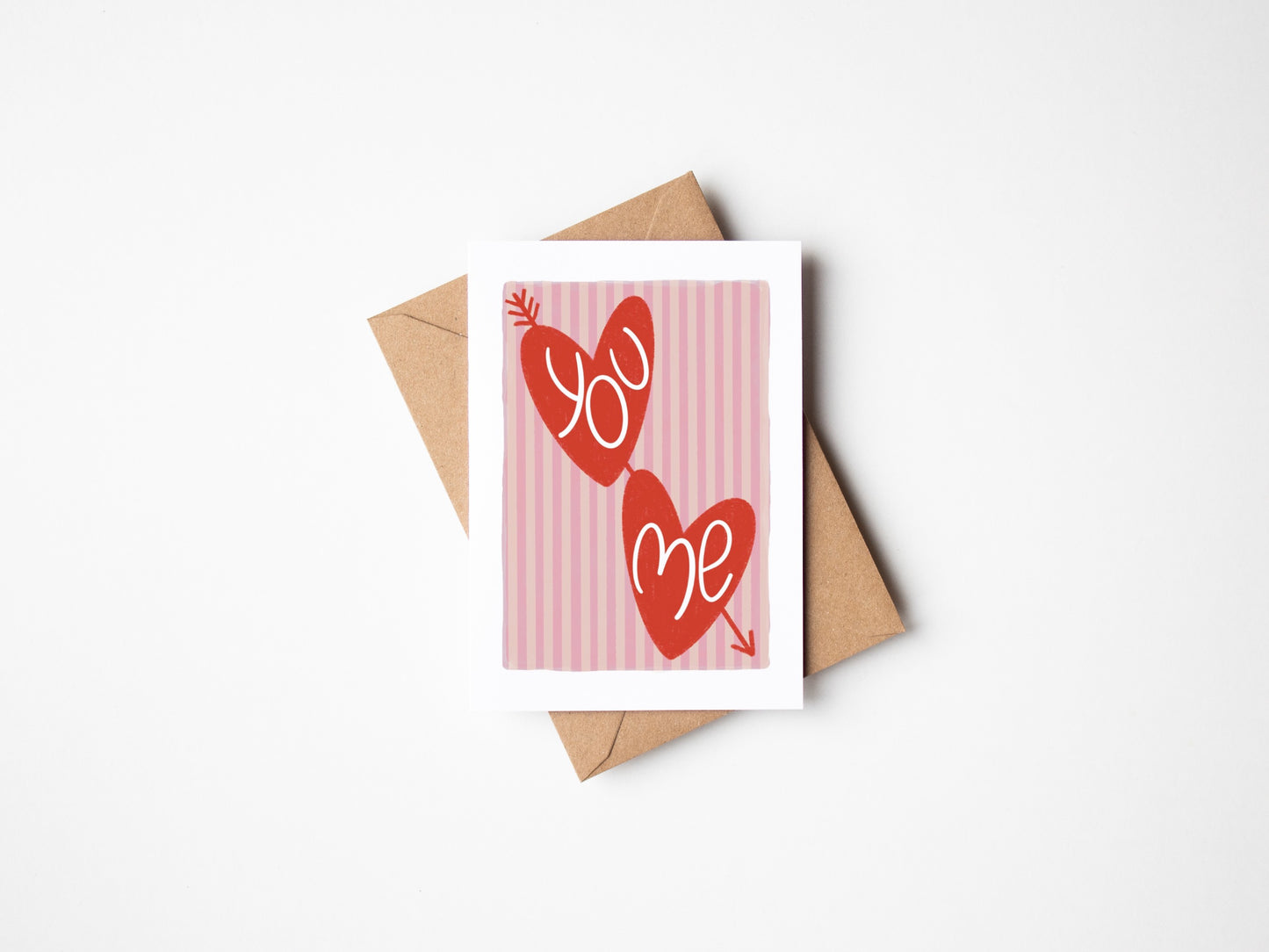 A6 white premium textured recyclable greeting card with light pink striped background, white border and two red love hearts with arrow through and the words ‘you’ and ‘me’ handwritten inside hearts. Brown Kraft envelope included.