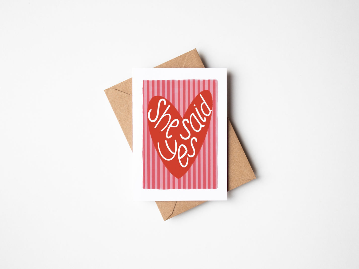 A6 premium textured card with white border, pink and red striped background and red love heart with the words “she said yes” handwritten inside heart. Brown Kraft envelope included.