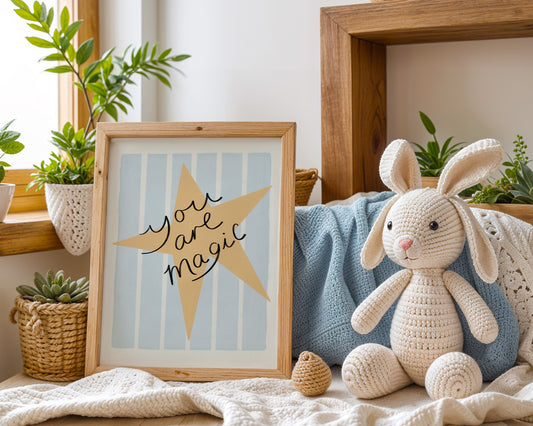 Framed artwork with a yellow star and 'You are magic' text on a light blue background, leaning against a white wall.