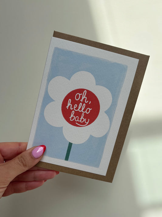 A6 premium textured recyclable card with white border, blue background and white daisy with red centre and the words ‘oh hello baby’ written inside centre of flower. Brown Kraft envelope included