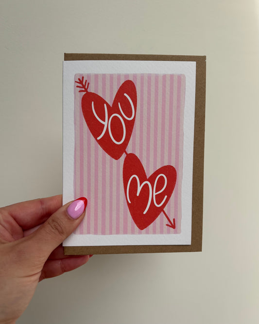 A6 white premium textured recyclable greeting card with light pink striped background, white border and two red love hearts with arrow through and the words ‘you’ and ‘me’ handwritten inside hearts. Brown Kraft envelope included.