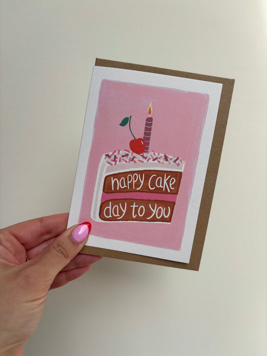 A6 premium textured recyclable greeting card with white border and pink background with drawing of a chocolate cake with white icing, pink and purple sprinkles, purple and white strped candle and cherry on top and the hand written word “happy cake day to you”. Brown Kraft envelope included.