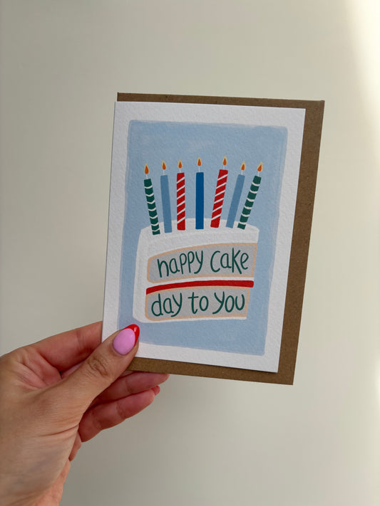 A6 premium textured recyclable greeting card with white border and blue background featuring a drawing of a slice of vanilla sponge cake with white icing and jam centre, topped with red, blue and green candles and the words “happy cake day to you”. Brown Kraft envelope Included.