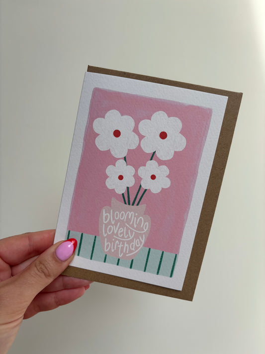 A6 premium textured recyclable greeting card with white border and pink background with beige vase on mint green striped tablecloth and bunch of white daises with red centres. Brown Kraft envelope included.