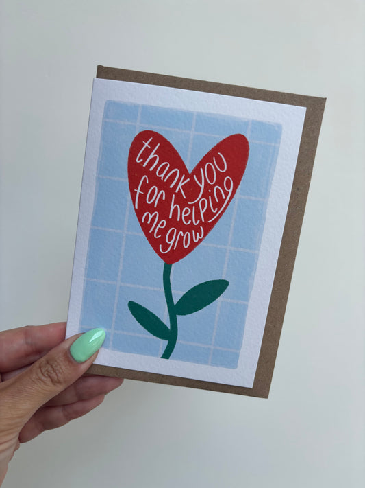 A6 premium textured recyclable teacher card with white border and blue and white grid background and red flower in a heart shape with the words “thank you for helping me grow” handwritten within it. Brown Kraft envelope included.