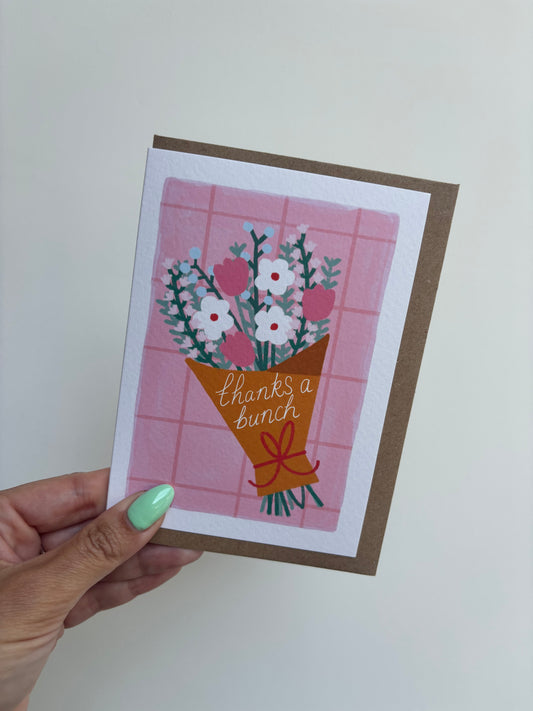 A6 recyclable greeting card with white border, pink grid background and a bunch o pastel flowers wrapped in brown paper tied with a red bow. Brown Kraft envelope included.