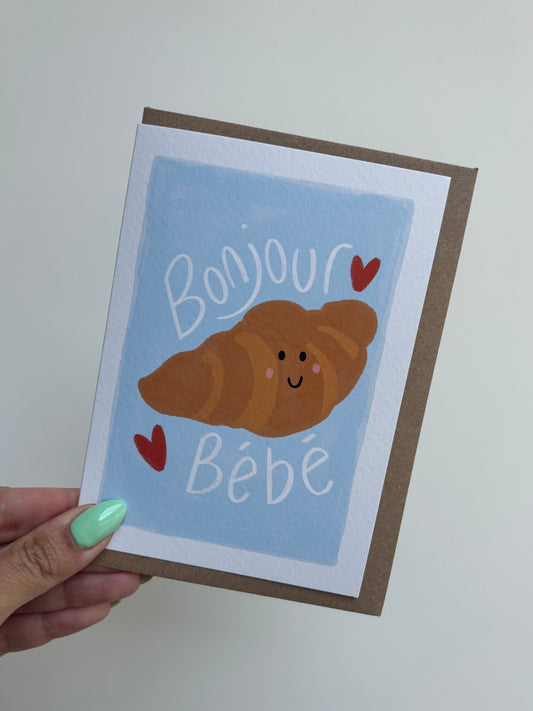 A6 premium textured recyclable greeting card with white border, blue background and croissant drawing with cute face and two love hearts with the words “bonjour Bebe”. Brown Kraft envelope included.