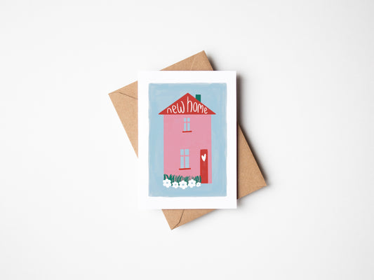 A6 white premium textured recyclable greeting card with white border, blue background and pink and red home with heart on the door and wording “new home” handwritten inside the roof of the house. Brown Kraft envelope included.