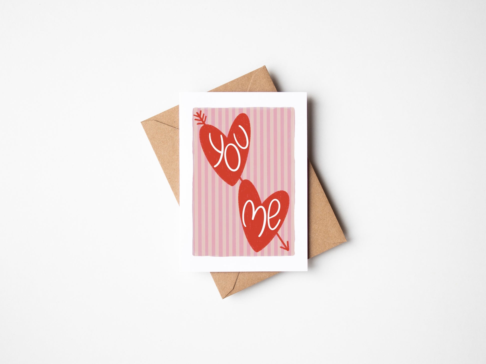 A6 white premium textured recyclable greeting card with light pink striped background, white border and two red love hearts with arrow through and the words ‘you’ and ‘me’ handwritten inside hearts. Brown Kraft envelope included.