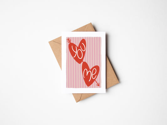 A6 white premium textured recyclable greeting card with light pink striped background, white border and two red love hearts with arrow through and the words ‘you’ and ‘me’ handwritten inside hearts. Brown Kraft envelope included.
