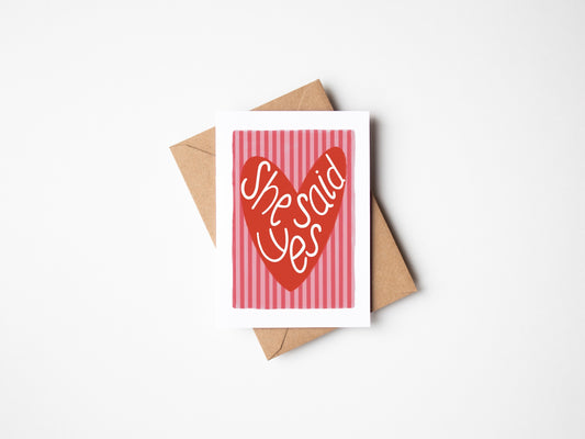 A6 premium textured card with white border, pink and red striped background and red love heart with the words “she said yes” handwritten inside heart. Brown Kraft envelope included. 