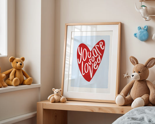 Kids wall art with white border, light blue background, red heart and the words ‘you are so loved’ in white hand written text.