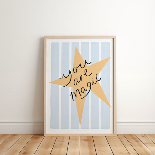 Framed artwork with a yellow star and 'You are magic' text on a light blue striped background, leaning against a white wall.