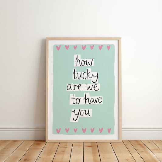 Framed print with 'how lucky are we to have you' text on a light green background with pink hearts, placed on a wooden floor.