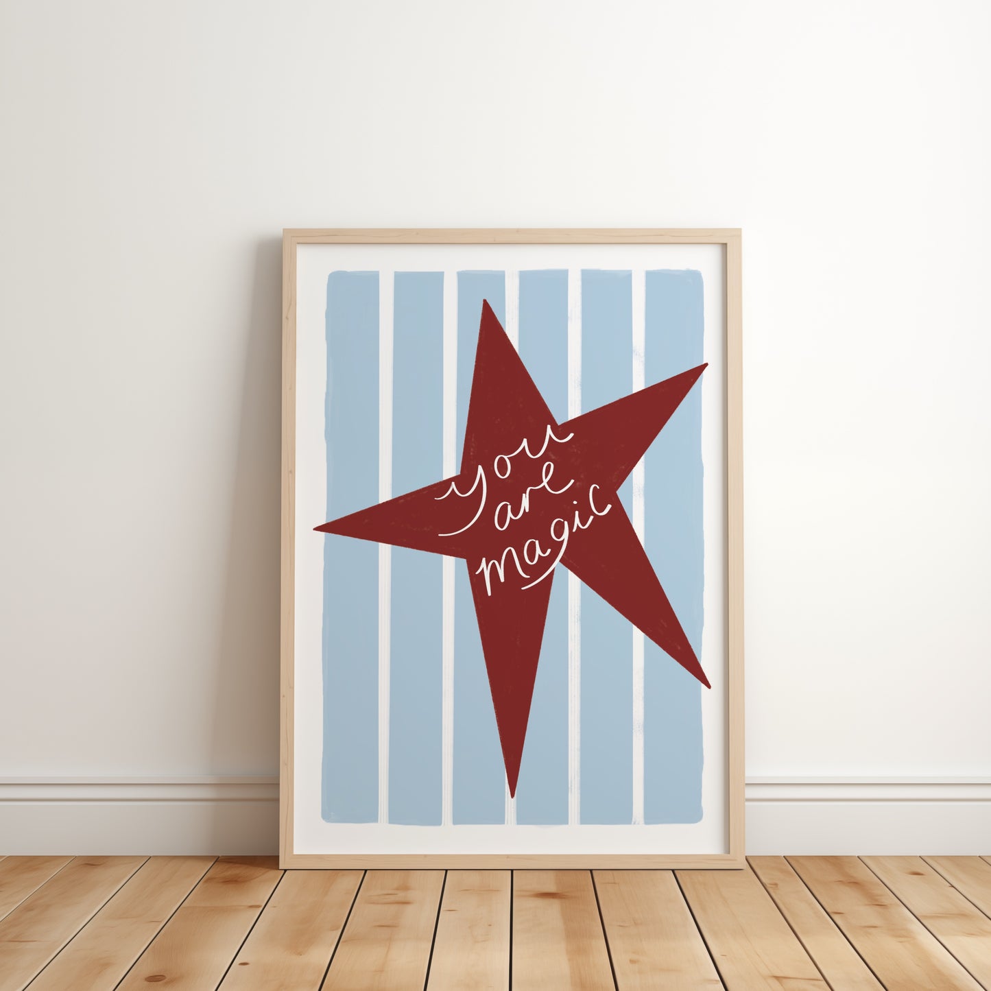Framed artwork with a red star and 'You are magic' text on a blue striped background, leaning against a white wall.