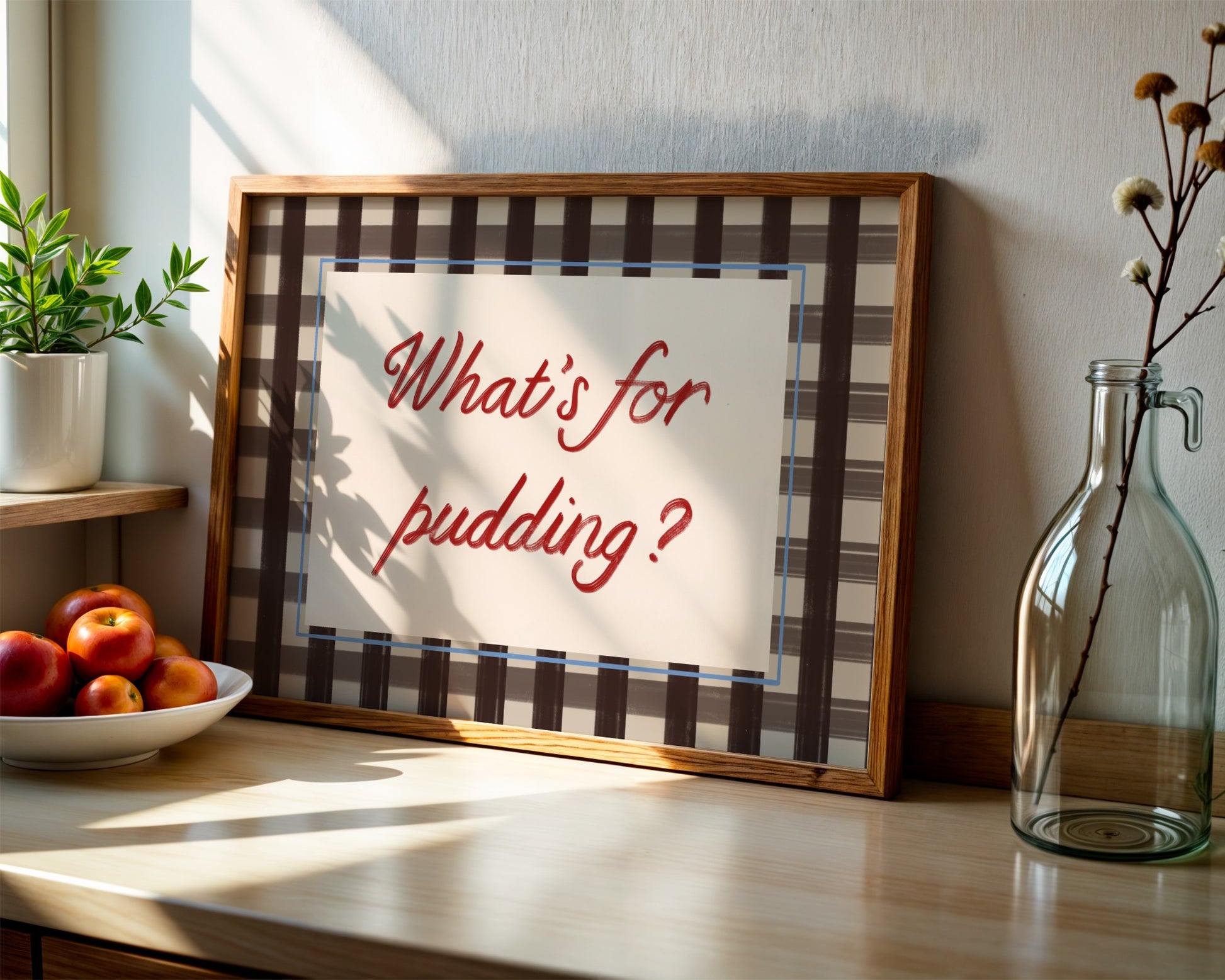 Framed sign with 'What's for pudding?' text on a kitchen counter with apples and a plant.
