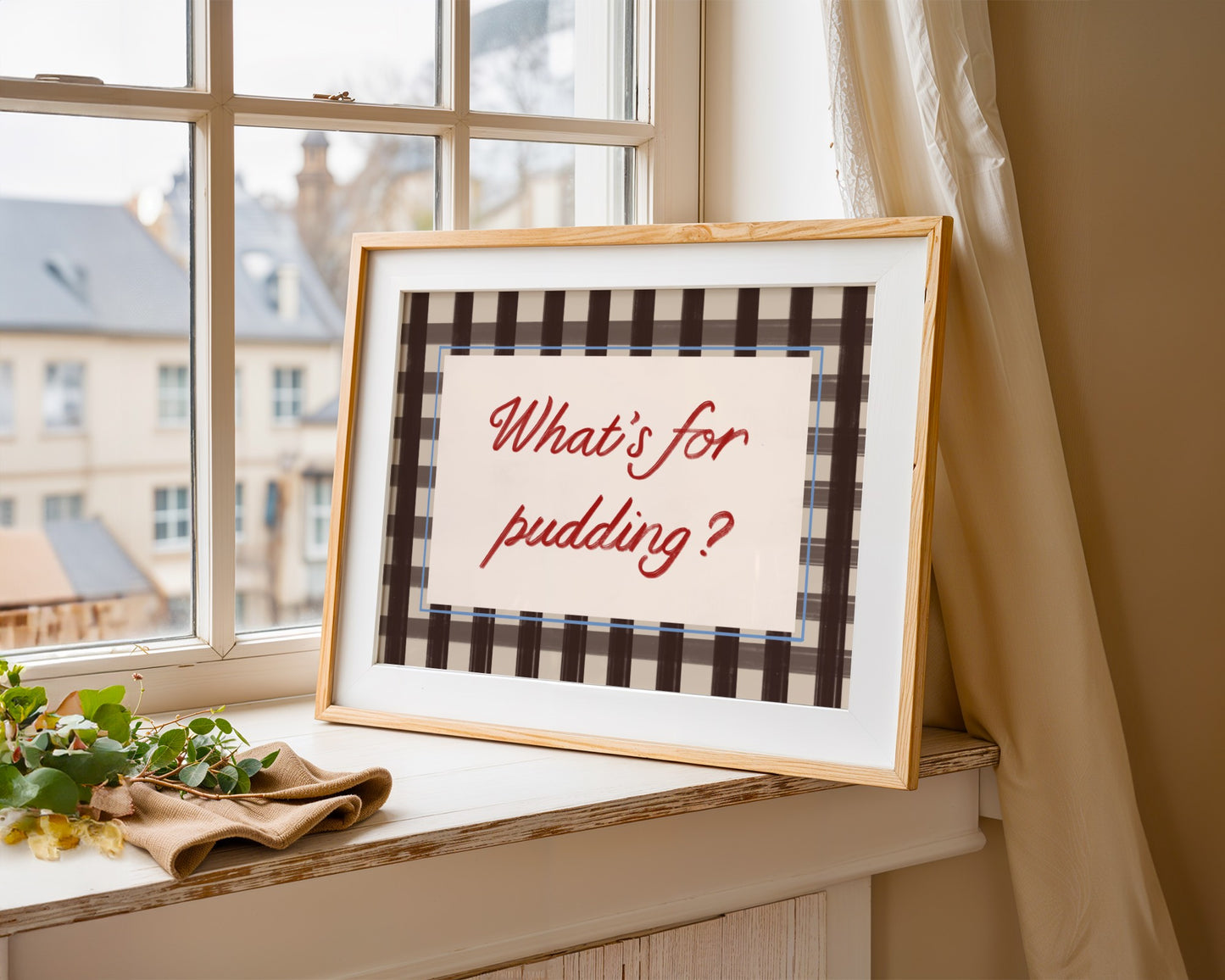 Framed picture with 'What's for pudding?' text on a windowsill with a view of buildings outside.