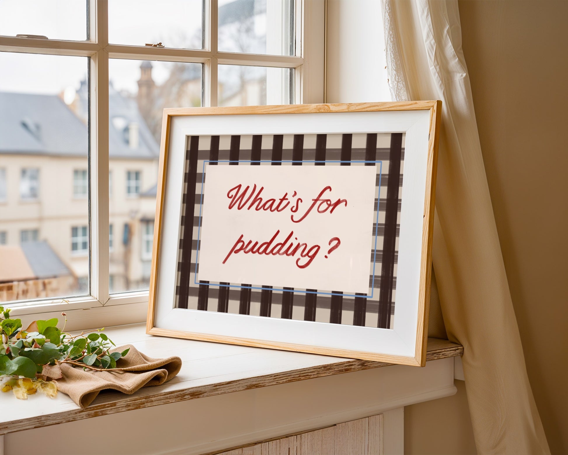 Framed picture with 'What's for pudding?' text on a windowsill with a view of buildings outside.