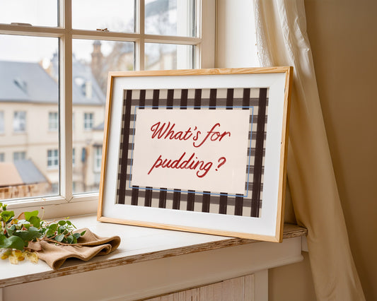 Framed picture with 'What's for pudding?' text on a windowsill with a view of buildings outside.