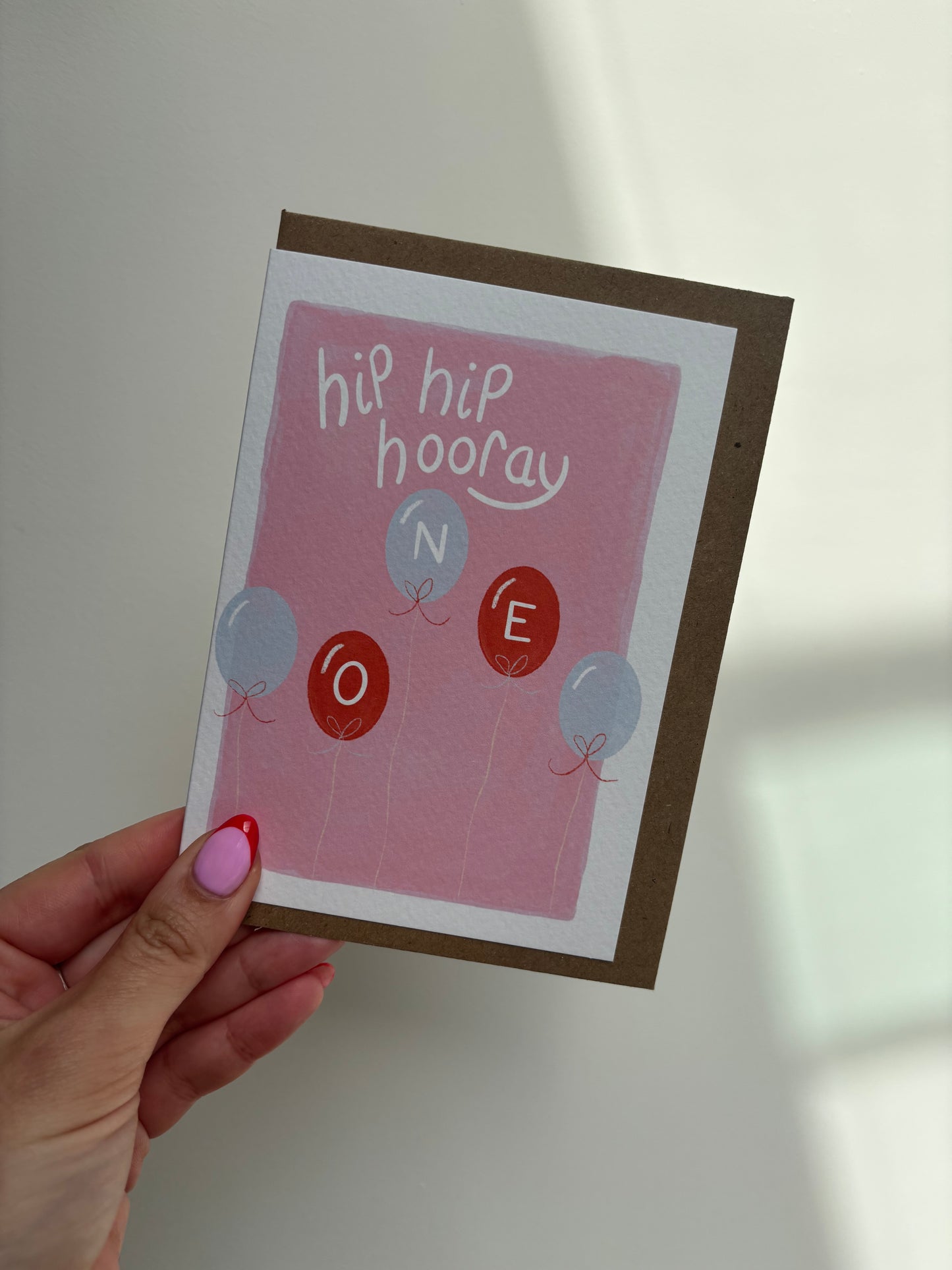 A6 premium textured recyclable greeting card with white border and pink background with five red and blue balloons with letters “ O N E” written inside them and the words “hip hip hooray” handwritten in white above. Brown Kraft envelope included.