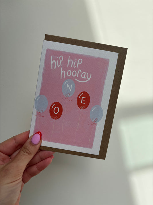 A6 premium textured recyclable greeting card with white border and pink background with five red and blue balloons with letters “ O N E” written inside them and the words “hip hip hooray” handwritten in white above. Brown Kraft envelope included.