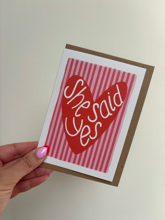 A6 premium textured card with white border, pink and red striped background and red love heart with the words “she said yes” handwritten inside heart. Brown Kraft envelope included. 