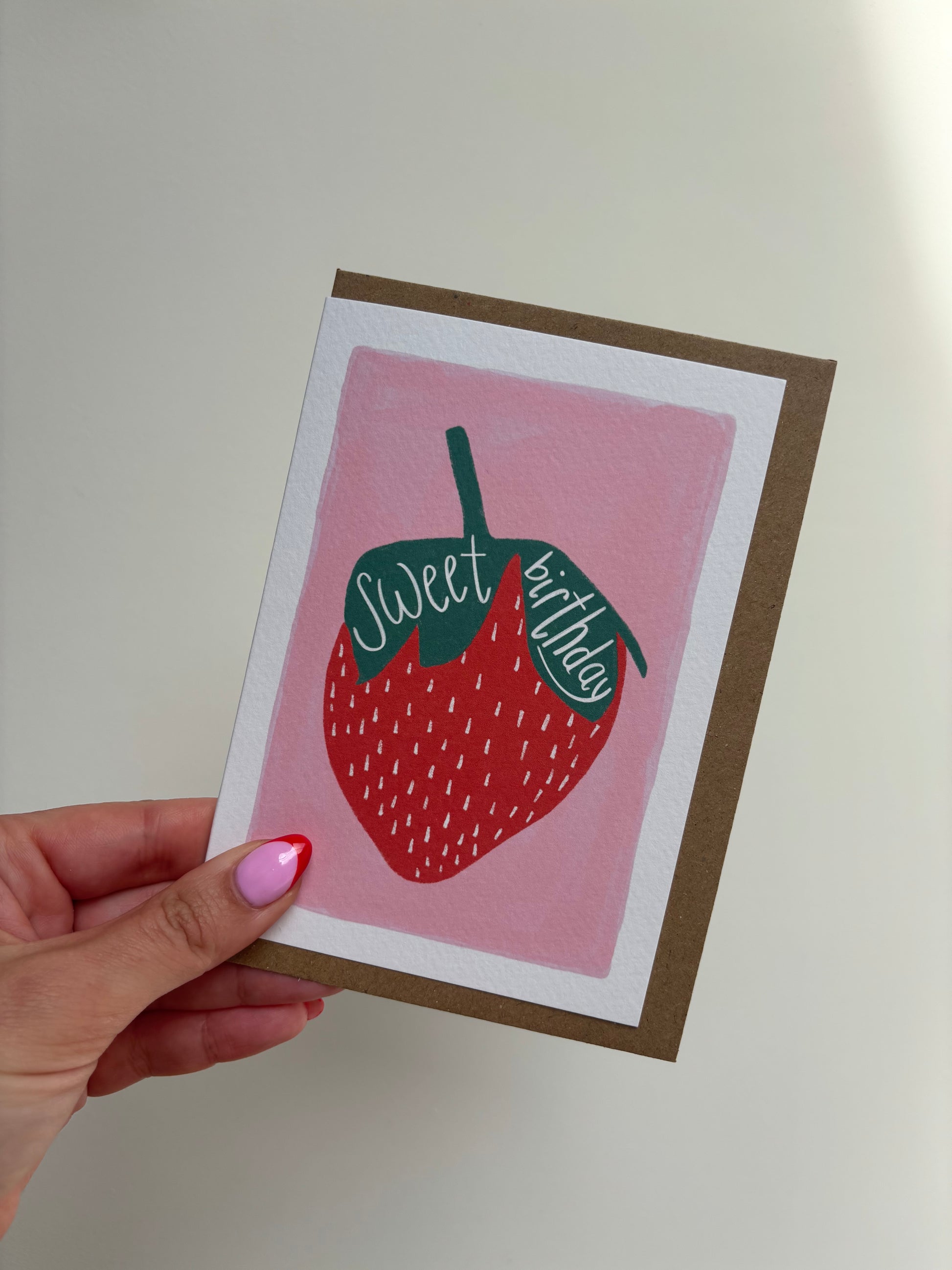 A6 premium textured recyclable greeting card with white border and pink background with strawberry drawing and the words “sweet birthday” handwritten inside leaves. Brown Kraft envelope included.