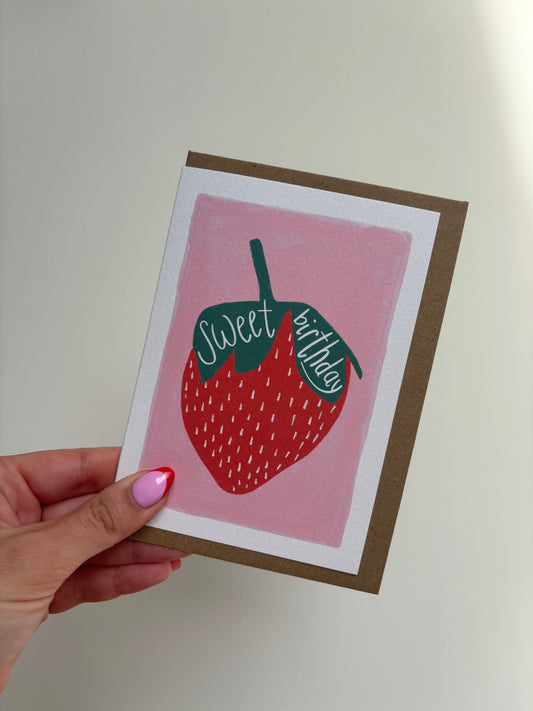 A6 premium textured recyclable greeting card with white border and pink background with strawberry drawing and the words “sweet birthday” handwritten inside leaves. Brown Kraft envelope included.