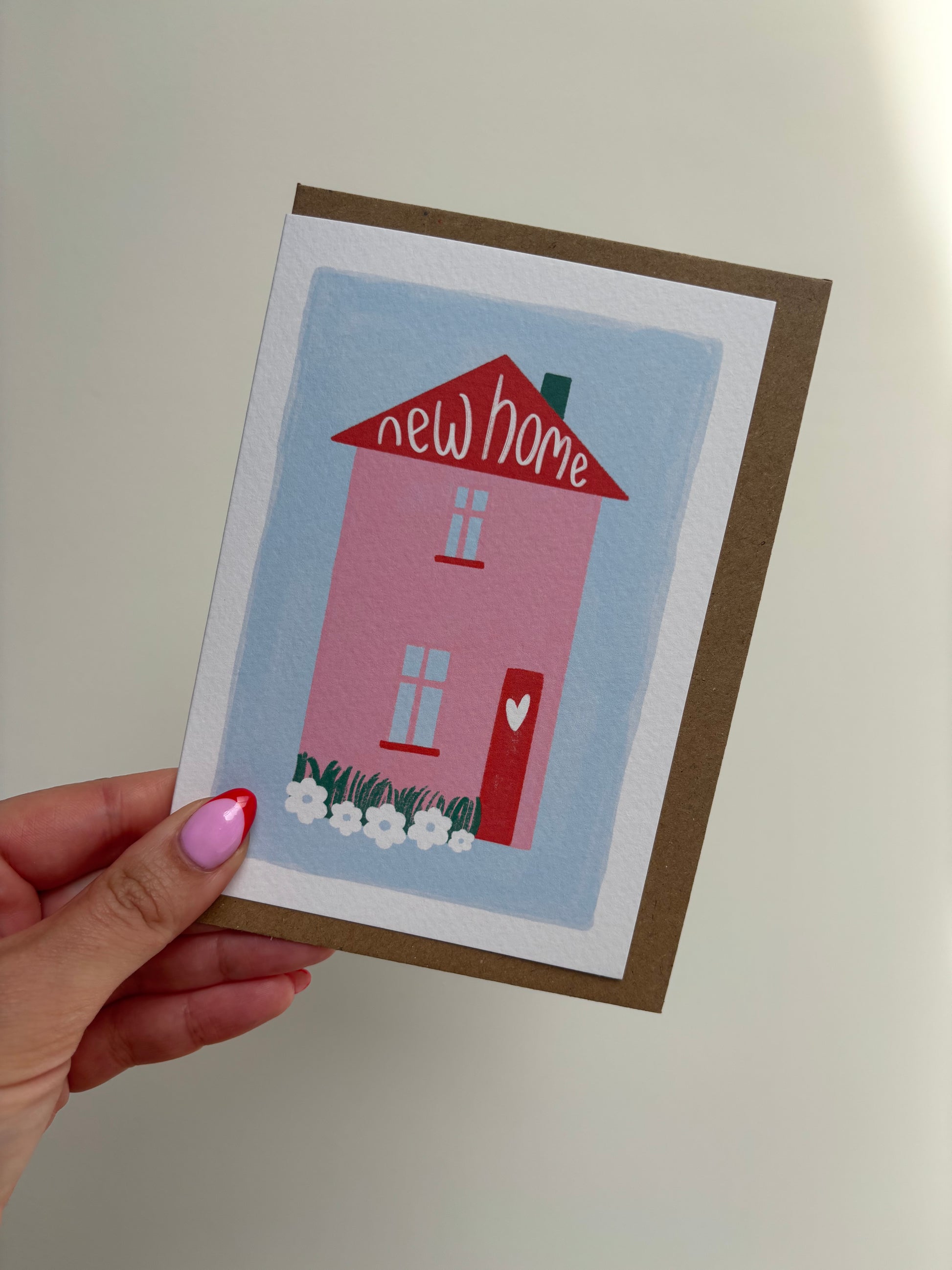 A6 white premium textured recyclable greeting card with white border, blue background and pink and red home with heart on the door and wording “new home” handwritten inside the roof of the house. Brown Kraft envelope included.