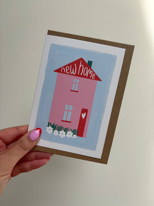 A6 white premium textured recyclable greeting card with white border, blue background and pink and red home with heart on the door and wording “new home” handwritten inside the roof of the house. Brown Kraft envelope included.