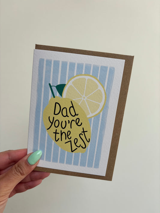 A6 white greeting card with blue and white striped background featuring a lemon and wording ‘Dad, you’re the zest’ . Features brown Kraft envelope.